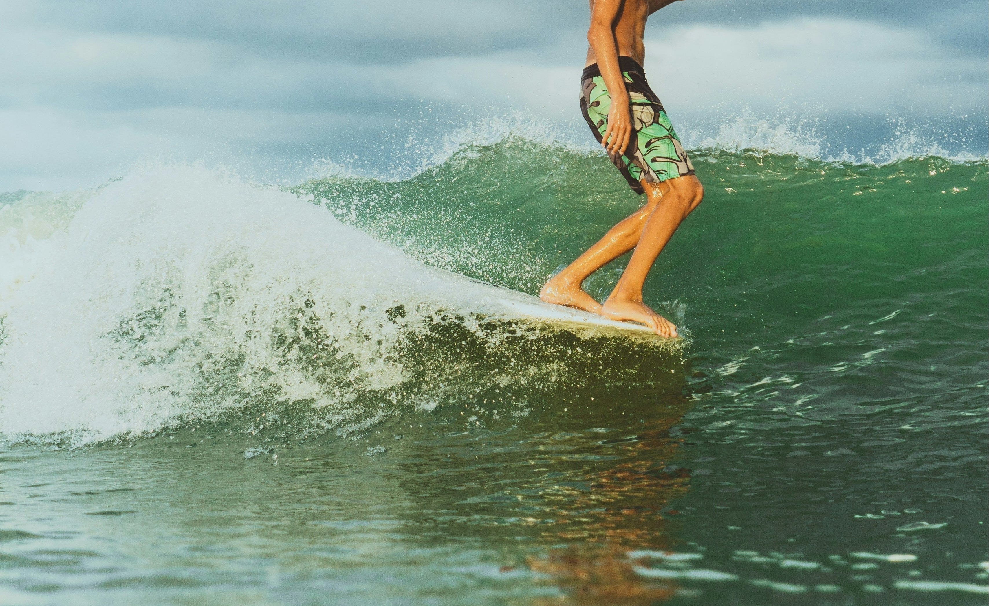 A man surfing on a green ocean wave under a cloudy sky, wearing green patterned swim trunks.